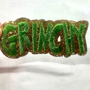 May include: Embroidered patch featuring the word "GRINCHY" in vibrant green thread. The letters are stitched onto a glittery, gold-toned base, creating a festive design. This patch is likely intended for application on clothing or accessories.