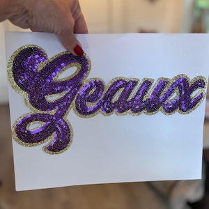 May include: A white paper with the word "Geaux" in purple sequins with a gold glitter outline. The word is in a cursive font. The paper is being held by a person.