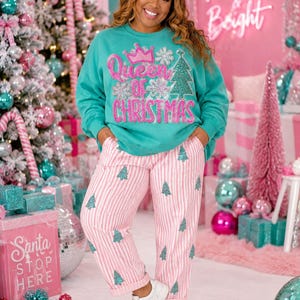 May include: A teal long-sleeve sweatshirt featuring a pink sequined crown and the text Queen of Christmas, paired with white and pink striped pants decorated with small green sequined Christmas trees and white sneakers with green star accents.