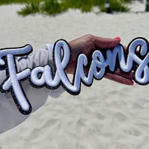 Può includere: Una toppa ricamata bianca con la scritta "Falcons" in corsivo. Le lettere sono bordate di nero con un bordo glitterato. La toppa è tenuta su uno sfondo di sabbia e vegetazione.