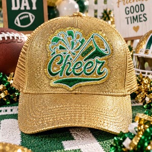 May include: A gold glitter baseball cap featuring a central embroidered patch with the word Cheer in green and white, accompanied by a graphic of a pom-pom and a megaphone. The cap has a structured front panel and a mesh back, with a shimmering finish.