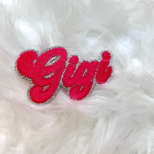 Puede incluir: Parche de fieltro rosa con el nombre "Gigi" en letras de purpurina plateada.