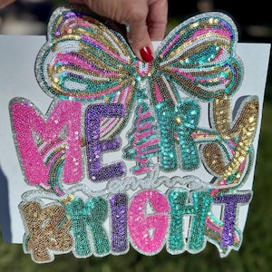 Puede incluir: Un letrero festivo con las palabras "MERRY and BRIGHT" en letras de lentejuelas. El letrero presenta un gran lazo y un diseño de árbol de Navidad, con una paleta de colores rosa, turquesa, dorado y morado. El letrero está sostenido por una mano.