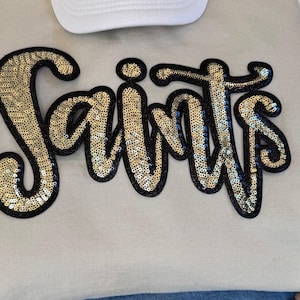 Toppa con scritta Saints in paillettes: applicazione termoadesiva con glitter neri, mascotte della scuola