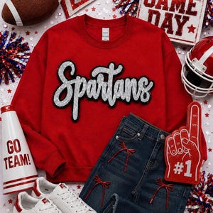 Puede incluir: Una sudadera roja de cuello redondo con la palabra Spartans en lentejuelas plateadas, vaqueros azul oscuro con lazos rojos, zapatillas blancas con estrellas rojas, un balón de fútbol americano, un megáfono blanco y un dedo de espuma rojo.