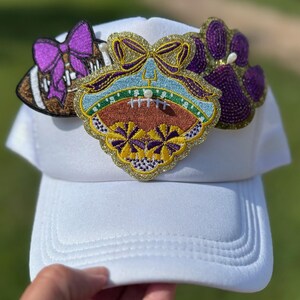 Puede incluir: Gorra de béisbol blanca adornada con tres parches decorativos. Los parches incluyen un balón de fútbol con un lazo morado, un emblema con temática de fútbol y una huella de pata morada. Los parches tienen detalles dorados y morados.