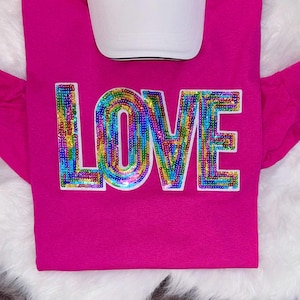May include: A hot pink shirt with the word "LOVE" spelled out in rainbow-colored sequins. A white baseball cap is placed above the shirt. The shirt is on a white, fluffy surface.