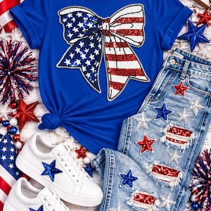 Op de afbeelding: Een koningsblauw t-shirt met een grote lovertjesstrik in Amerikaanse vlagpatronen, gecombineerd met een lichtblauwe, versleten spijkerbroek met lovertjessterren en vlagpatches, en witte sneakers met blauwe lovertjessterren.