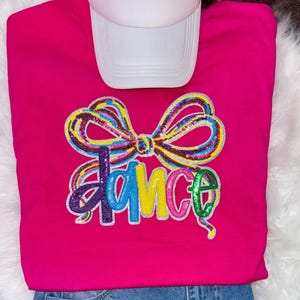 Puede incluir: Una sudadera rosa intenso con la palabra "DANCE" en letras de lentejuelas coloridas y un lazo de lentejuelas. Una gorra de béisbol blanca descansa encima. La sudadera se combina con mezclilla azul.