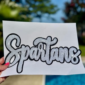 May include: A white patch featuring the word Spartans written in a cursive script. The lettering is filled with white sequins and outlined with a thick black border, creating a high-contrast design on a plain white rectangular background.