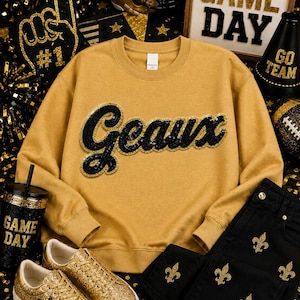 May include: A gold-colored crewneck sweatshirt featuring the word Geaux in black sequins with gold trim. The sweatshirt is paired with black jeans decorated with gold fleur-de-lis patterns and gold glitter sneakers with a black star detail.
