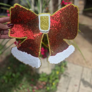 Puede incluir: Un gran lazo rojo y dorado con lentejuelas y ribete de piel blanca. El lazo tiene forma de campana de Navidad.