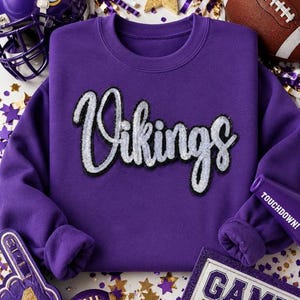 May include: A purple crewneck sweatshirt featuring the word Vikings in large, silver sequined script lettering across the chest. The garment has long sleeves and a ribbed neckline, cuffs, and hem, constructed from a solid purple fabric.