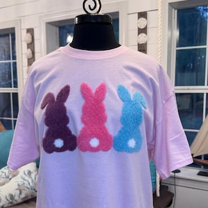 Könnte beinhalten: Hellrosa T-Shirt mit drei flauschigen Hasen-Silhouetten in Lila, Rosa und Blau, jeweils mit weißem Schwanz. Das Shirt wird auf einem schwarzen Schaufensterpuppenkopf präsentiert.
