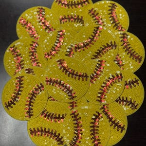 Peut inclure: Une collection d'écussons jaunes en forme de balle de softball, ornés de paillettes. Les écussons présentent des coutures en paillettes marron et rouges, imitant les coutures d'une balle de softball. Les écussons sont disposés en cercle.