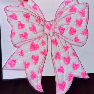 May include: A large, decorative bow with a white background and a pink outline. The bow is adorned with numerous pink heart-shaped sequins. The bow is held by a hand with red nail polish.