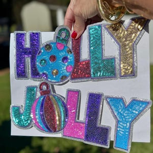 May include: Two sequined signs spelling "HOLY JOLLY". The top sign has a purple "H", a red "O", a teal "L", and a gold "Y". The bottom sign has a teal "J", a pink "O", a purple "L", and a blue "Y". Each letter has a silver border.