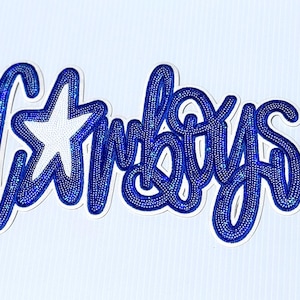 May include: Blue rhinestone-studded Cowboys script with a white star, set against a light blue background. The word "Cowboys" is written in a cursive font, with each letter outlined in rhinestones.