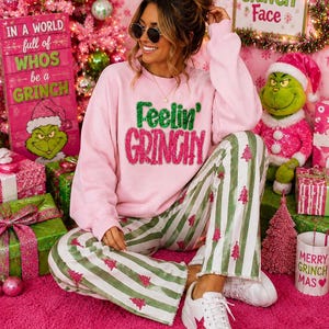 May include: A light pink long-sleeve sweatshirt with the text Feelin' Grinchy in green and pink sequins, paired with white and green striped pants featuring small pink Christmas tree patterns and white sneakers with a pink star detail.