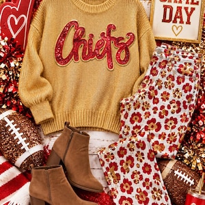May include: A gold knit sweater with the word Chiefs in red sequins, paired with white jeans featuring a red and gold floral pattern and brown ankle boots. A sign reads GAME DAY with a heart, alongside football-themed decor and pom-poms.