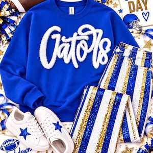 May include: A royal blue crewneck sweatshirt with white fuzzy chenille lettering that reads Gators, paired with white, royal blue, and gold sequined striped pants and white sneakers featuring a blue star on the side of each shoe.