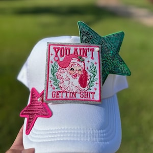 May include: White trucker hat with a Santa Claus patch that reads "YOU AIN'T GETTIN SHIT." The hat is decorated with a pink and a green star. The Santa patch has a pink border and a Santa Claus illustration.