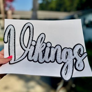 May include: A white sequin patch featuring the word Vikings in a cursive script font. The lettering is outlined in a thick black border, creating a high-contrast design. The patch is displayed on a plain white background, showcasing the texture.