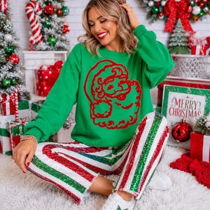 May include: A bright green long-sleeve sweatshirt featuring a large red graphic of Santa Claus on the front, paired with white, red, and green sequined striped pants and white sneakers with a green star detail on the side.