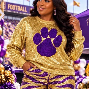 May include: A long-sleeved gold sequin sweatshirt featuring a large purple paw print graphic on the chest, paired with matching gold and purple zebra-striped sequin pants. The outfit has a crew neckline and ribbed cuffs at the wrists and waist.