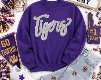 May include: A purple crewneck sweatshirt with the word Tigers in silver sequins, light wash denim jeans with tiger face patches, white sneakers with star accents, a purple football helmet, a football, and a Game Day sign.