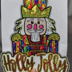 May include: A sequined Christmas-themed patch featuring a cartoon nutcracker soldier with a gold crown and a white beard. The nutcracker's uniform is a rainbow of colors. The text "Holly Jolly" is below the nutcracker.