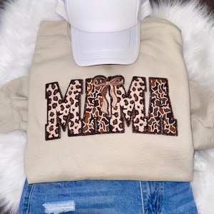 Op de afbeelding: Een beige sweatshirt met het woord "MAMA" geborduurd in een luipaardprint. Een witte truckerpet ligt erop. Het sweatshirt wordt gecombineerd met een blauwe spijkerbroek.