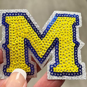 May include: A letter M patch featuring a silver glitter base, a border of blue sequins, and a center filled with yellow sequins. The letter has a block-style font design with sharp edges and a textured, sparkling appearance throughout the surface.
