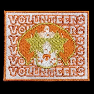 Può includere: Una toppa ricamata bianca con la parola "VOLUNTEERS" ripetuta in un motivo circolare. Al centro c'è un cane in stile cartone animato che indossa occhiali da sole con un design a forma di stella.