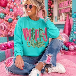May include: A bright turquoise long-sleeve sweatshirt featuring the text Merry Christmas in large, glittery pink and green sequined letters. The sweatshirt is paired with blue denim jeans that have distressed patches revealing pink sequin fabric underneath.