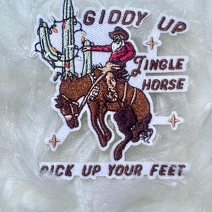 May include: Embroidered patch featuring a cowboy riding a bucking horse, with a cactus decorated with Christmas lights. Text includes "Giddy Up," "Jingle Horse," and "Pick Up Your Feet." The cowboy wears a red shirt and brown hat.