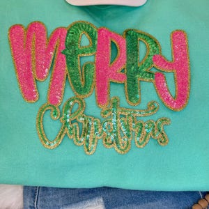 K&ouml;nnte beinhalten: Ein mintgr&uuml;nes Sweatshirt mit den Worten "MERRY CHRISTMAS" in Paillettenbuchstaben. Das Wort "MERRY" ist in rosa und gr&uuml;nen Pailletten mit goldenem Rand, und "CHRISTMAS" ist in gr&uuml;nen Pailletten mit goldenem Rand. Oben ist eine wei&szlig;e Baseballkappe zu sehen.