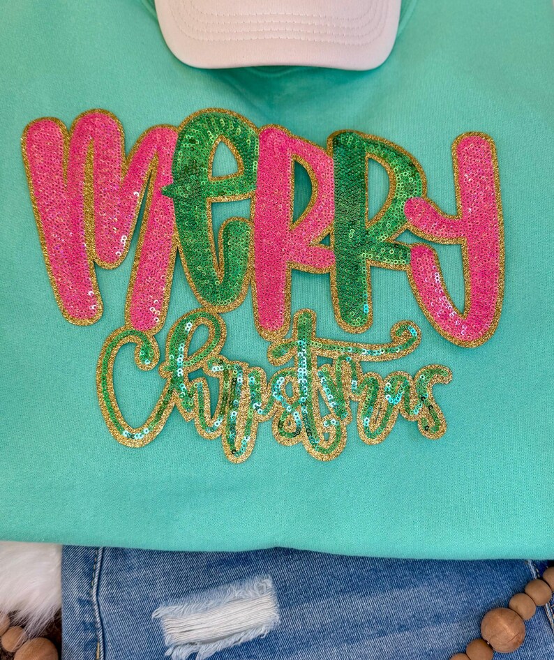 K&ouml;nnte beinhalten: Ein mintgr&uuml;nes Sweatshirt mit den Worten "MERRY CHRISTMAS" in Paillettenbuchstaben. Das Wort "MERRY" ist in rosa und gr&uuml;nen Pailletten mit goldenem Rand, und "CHRISTMAS" ist in gr&uuml;nen Pailletten mit goldenem Rand. Oben ist eine wei&szlig;e Baseballkappe zu sehen.