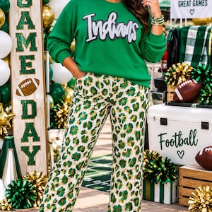 May include: A bright green long-sleeve sweatshirt with the word Indians in white and black script across the chest, paired with white pants featuring a green and gold leopard print pattern and white sneakers with a green star detail.