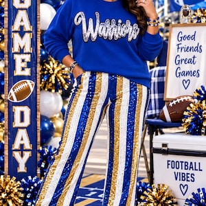 May include: A royal blue long-sleeve sweatshirt featuring the word Warriors in silver sequins across the chest, paired with high-waisted trousers decorated in vertical stripes of shimmering blue, gold, and white sequins, worn with white boots.