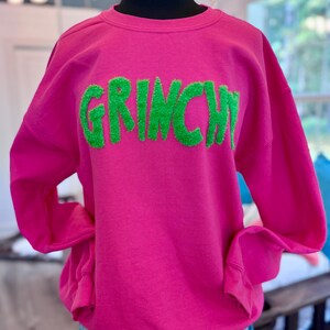 May include: A bright pink sweatshirt with the word "GRINCH" in green, textured lettering. The long-sleeved top is displayed on a mannequin, paired with blue denim shorts. The sweatshirt is a casual, festive garment.