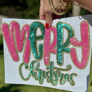 Puede incluir: Un letrero blanco con las palabras "MERRY Christmas" en letras de lentejuelas. La palabra "MERRY" está en lentejuelas rosas con ribete dorado, y la palabra "Christmas" está en lentejuelas verdes con ribete dorado.