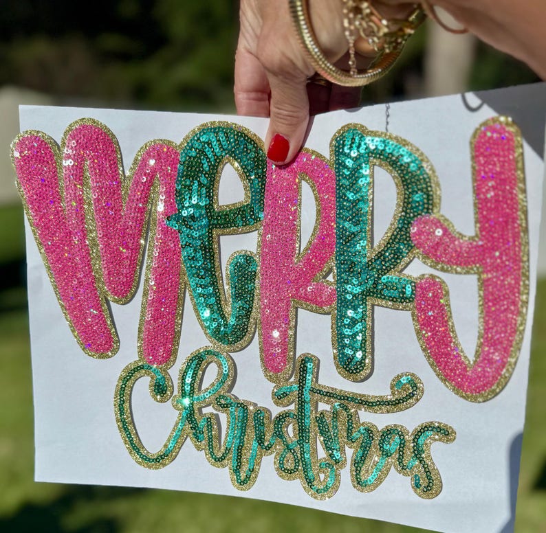 K&ouml;nnte beinhalten: Ein wei&szlig;es Schild mit den Worten "MERRY Christmas" in Paillettenbuchstaben. Das Wort "MERRY" ist in rosa Pailletten mit goldenem Rand gehalten, das Wort "Christmas" in gr&uuml;nen Pailletten mit goldenem Rand.