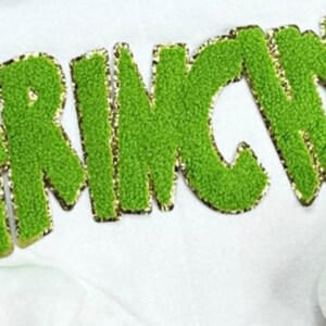 May include: A close-up of the word "GRINCH" in green, textured, embroidered letters with a gold border. The letters are on a white background, likely a piece of clothing or fabric.