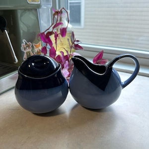 May include: A blue and white ceramic sugar bowl with a lid and a matching creamer. The sugar bowl is round with a dark blue lid. The creamer has a handle and a spout. The set is on a light-colored countertop.