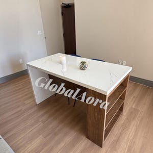 May include: A rectangular kitchen island or desk featuring a white marble-patterned countertop with grey veining. The base is constructed from brown wood, incorporating three open storage shelves on one side and a solid panel on the other.