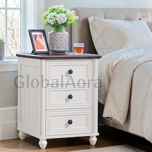 May include: A white nightstand with three drawers and a dark brown top. The nightstand has black knobs and is next to a bed with a beige headboard. A vase of flowers, a framed picture, and a glass of liquid sit on top.