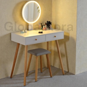 May include: A white dressing table with two drawers and wooden legs, accompanied by a grey stool. A circular mirror with a lit edge is above the table. Makeup and accessories are on the tabletop.