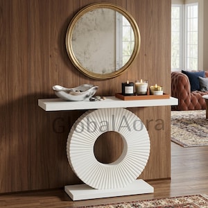 May include: A white console table featuring a large, circular, ribbed base and a rectangular top. On the table sits a silver, irregular-shaped bowl and a rectangular wooden tray holding three cylindrical candles of varying heights.