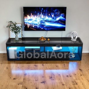 May include: A black entertainment unit with blue LED lighting, a large flat-screen TV, and the text "GlobalAora". The unit has open shelving and a glass top. Decorative items include a plant, fruit, and headphones.